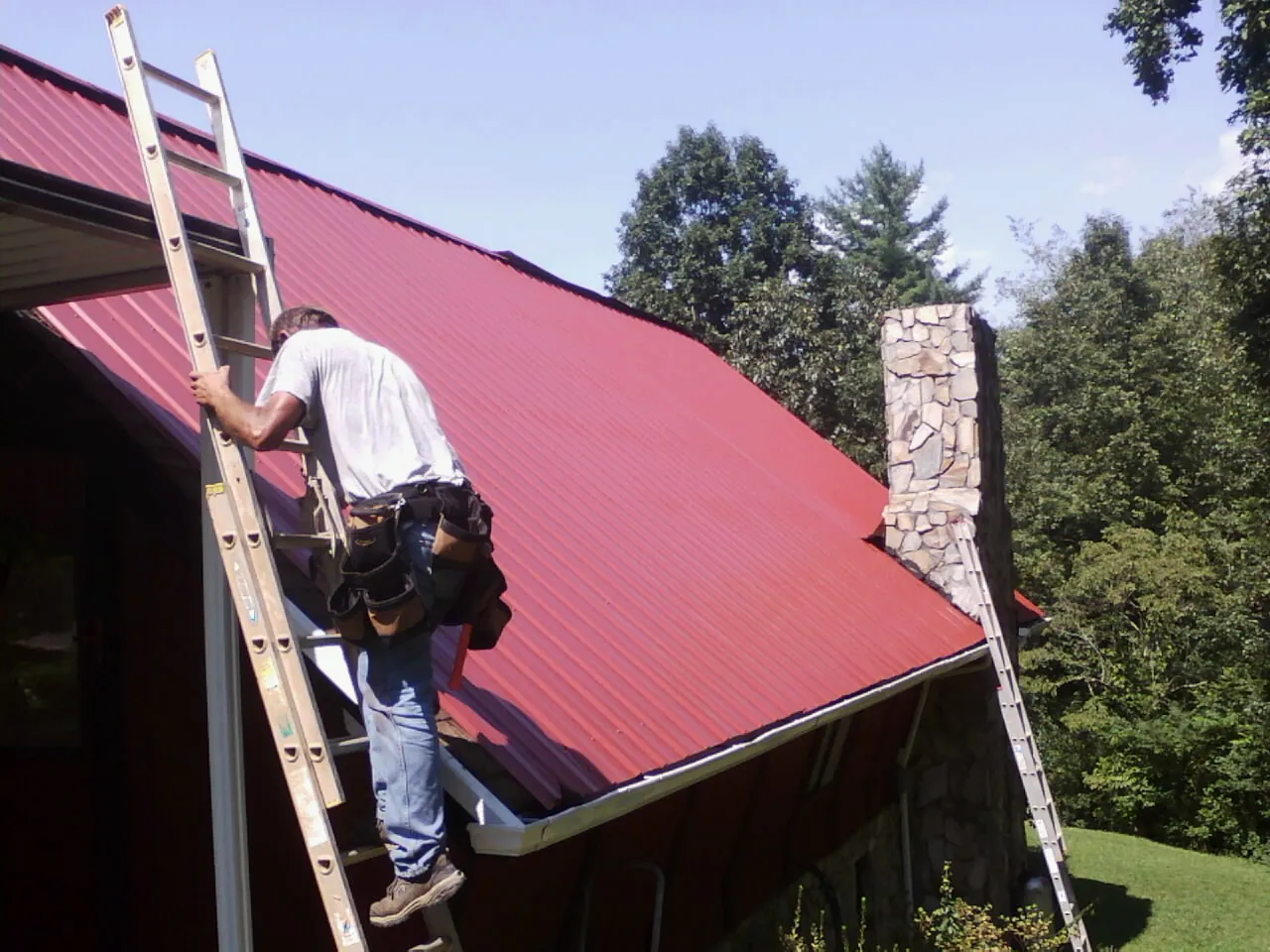 Roofer on a ladder beside a red metal roof and stone chimney — Soffit & Fascia Repair by Roofing Brothers in Garwood